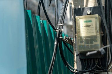Kablolar ve yeşil çitin yanındaki elektrik sayacının yakın görüntüsü. Bu resim kentsel bir ortamda temel kullanım altyapısını yakalar.