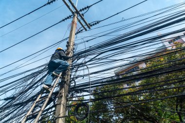 Bir işçi, kentsel bir ortamda karmaşık bir kablo dizisini yönetmek için elektrik bakım ve güvenliğinin temel yönlerini vurgulamak için elektrik direğine tırmanır..