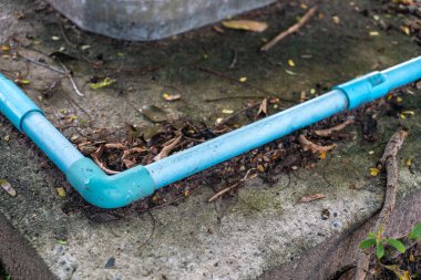 Beton bir yüzeyde duran mavi plastik borunun yakın görüntüsü. Çevreleyen elementler arasında toprak ve yapraklar, açık hava ortamını betimleyen.