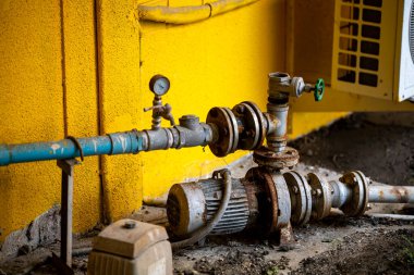 Basınç göstergesi ve motoru olan endüstriyel bir boru sisteminin ayrıntılı görüntüsü, canlı sarı bir duvara karşı kurulmuş, kentsel bir ortamda araçlar ve makineler ön plana çıkarılıyor..