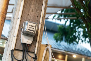 Beton bir duvara monte edilmiş metalik bir elektrik sayacının yakınlaştırılması, kentsel bir ortamda kullanım ve ölçüm sistemlerini gösteren kablolar ve bağlantılar sergiliyor.