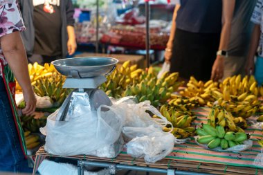 Kalabalık bir markette satılık taze muz manzarası. Bir satıcı, renkli ürünlerle çevrili bir ölçekte muzları tartar. Toplumu ve yerel lezzeti ön plana çıkarır..