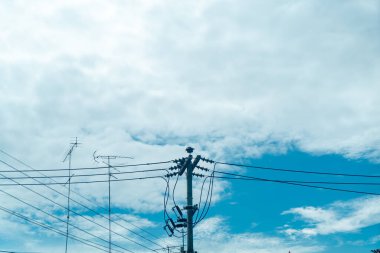 Yumuşak bulutlarla dolu parlak mavi gökyüzüne uzanan elektrik hatlarının huzurlu görüntüsü. Enerji altyapısını ve kentsel manzaraları resmetmek için ideal.