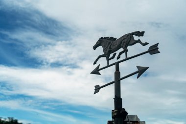 Çarpıcı bir metal rüzgar gülü dörtnala giden bir atı dinamik bulutlara karşı sergiliyor. Yön ve doğa temaları için ideal..