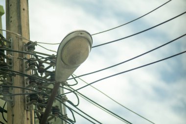 A close-up of a streetlight mounted on a utility pole, surrounded by a network of power lines under a cloudy sky, illuminating the urban environment below.
