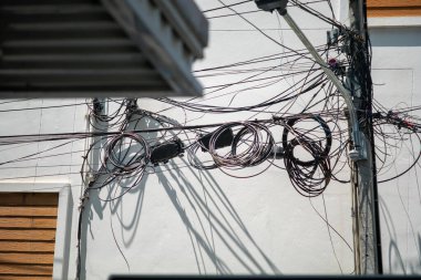 A tangled array of power lines casts intricate shadows against a building's white wall in an urban setting. The chaotic arrangement creates a striking visual contrast.
