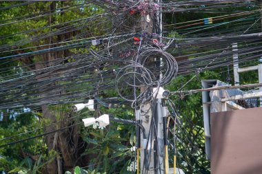 A detailed view of complex overhead electric wires intertwining with surveillance cameras, showcasing urban infrastructure against a backdrop of greenery.