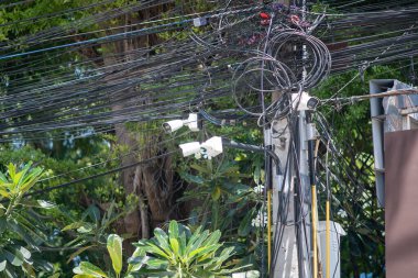 A detailed view of a utility pole showcasing a complex network of tangled electric wires and surveillance cameras among lush green foliage, highlighting urban infrastructure.