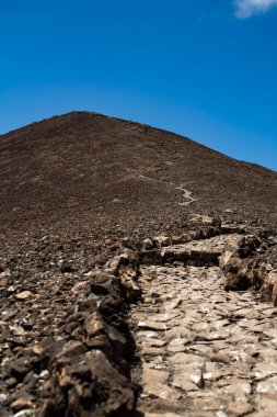 Sivri lav taşlarıyla tanımlanmış çorak bir volkanik yamacı saran dik bir patika. Bu ham arazi, dayanıklılık ruhunu ve hayattaki sembolik yokuş savaşlarını yakalar..