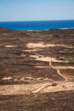 Kıvrımlı bir patika, ufuktaki mavi okyanusa doğru uzanan kuru volkanik araziyi kesiyor. Dönen yollar, hayatın kıvrımlarını, seçimlerini ve görünmeyen hedefleri temsil eder..