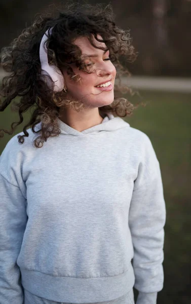 公園で夜に音楽を聴くヘッドフォンを身に着けている灰色のスポーツパーカーの若い美しい女性 — ストック写真