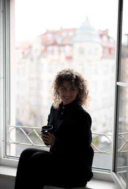 Kıvırcık saçlı kadın açık pencerenin yanında kahve içiyor.