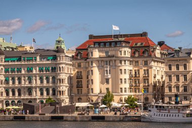 İsveç, Stockholm - 17 Temmuz 2022 Skeppsbron rıhtımından görülen Grand Hotel 'in kaplıca ve spor salonu bej rengi tarihi cephesi. Üstte beyaz bayrak, rıhtımda yayalar. Mavi bulut