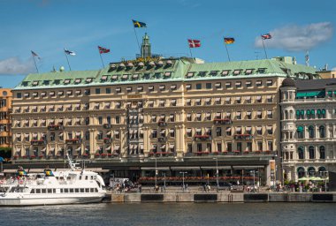 İsveç, Stockholm - 17 Temmuz 2022: Skeppsbron rıhtımından görünen Grand Hotel 'in ana binası, üzerinde ulusal bayraklar, rıhtımda yayalar ve mavi gökyüzü altında beyaz tekne