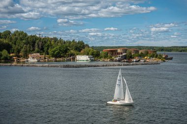 İsveç, Stockholm - 17 Temmuz 2022: Stora Hoggarn adasının güneyinde, kahverengi silindirik tankların bitişiğindeki barajı koruyan beyaz yelkenli yat