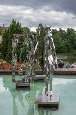 Finlandiya, Kotka - 18 Temmuz 2022: Sopokanlahti park ve gölü. Parlak metal modern sanat heykelleri yeşil su havuzunda. Gri bulutlar altında yeşil kuşak. Kırmızı çiçekler renk ekler