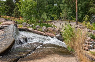 Finlandiya, Kotka - 18 Temmuz 2022: Sopokanlahti park ve gölü. Şelalenin üst kısmı, gri ve kahverengi yüzey ve beyaz köpüklü su. Arkaplan olarak yeşil yapraklar.