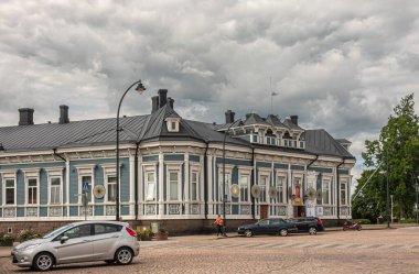 Finlandiya, Hamina - 18 Temmuz 2022: Belediye binasının karşısındaki tarihi mavi-beyaz ticari bina, önünde arabalar ve insanlar olan gri bulutların altında. Kültür merkezi dahil