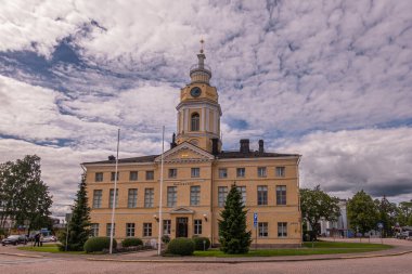 Finlandiya, Hamina - 18 Temmuz 2022: Yellow Stone kasabası veya belediye binasının arka tarafı, Haminan raatihuone, saat kulesi bu gri bulutların altında. Yeşil çimenler ve öndeki insanlar