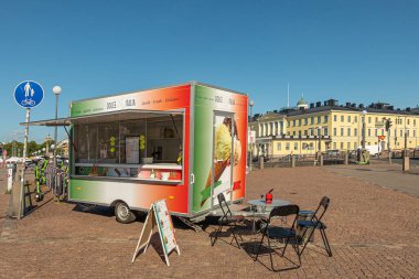 Helsinki, Finlandiya - 19 Temmuz 2022: Mavi gökyüzünün altındaki Kanavakadunsilta köprüsü yakınlarındaki meydanda renkli dondurma karavanı Dolce Italia. Masa ve sandalyeler. Başkanlık Sarayı arkada.