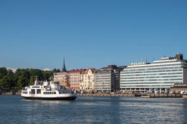 Helsinki, Finlandiya - 19 Temmuz 2022: Siyah-beyaz Suokki feribot feribotu arkada Etelaeranta caddesinin ön cepheleriyle mavi gökyüzünün altında limandan ayrılıyor. Palace Otel baskın. Park ve oyun alanı yeşil yapraklar