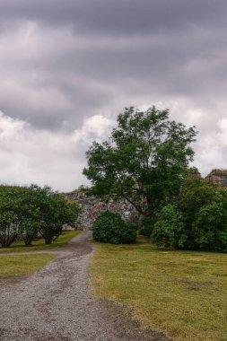 Helsinki, Finlandiya - 19 Temmuz 2022: Suomenlinna Kalesi. Gri çakıllı yol gri-kahverengi taşlı surlara çıkıyor. Şiddetli yağmurlu, karanlık bulutların altında yeşil ağaçlar arasında.