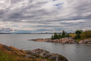 Helsinki, Finlandiya - 19 Temmuz 2022: Suomenlinna Kalesi. Gri bulutlar altında sanayi ve liman şehri Munkkisaari ile SW sahilindeki Deniz Koyu 'nda geniş bir manzara. Önde yeşil yapraklar var.