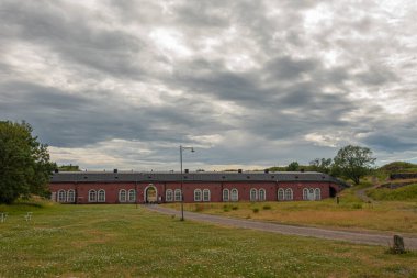 Helsinki, Finlandiya - 19 Temmuz 2022: Suomenlinna Kalesi. Askerler için uzun, kırmızı, tarihi taş bina, bugün Kuzey Kültür Merkezi, HIAP, gri bulutların altında, sarı çayırlarda geçiyor.