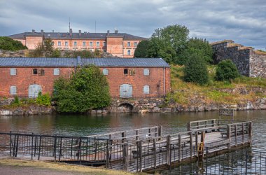 Helsinki, Finlandiya - 19 Temmuz 2022: Suomenlinna Kalesi. Doğu adasının tepelerinde, mavi bulutların altında büyük kırmızı binalar. Ön tarafta ahşap iskeleli deniz suyu. Tepede yeşil yapraklar