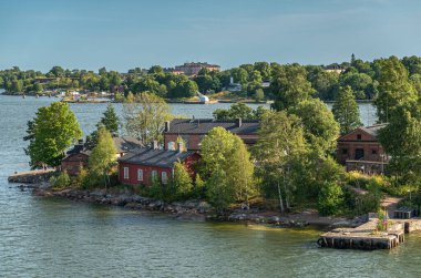 Helsinki, Finlandiya - 20 Temmuz 2022: Suomenlinna Kalesi. Mavi gökyüzünün altındaki kıyı şeridindeki kısa rıhtımların yakınındaki depolar. Bir sürü yeşil yeşillik..