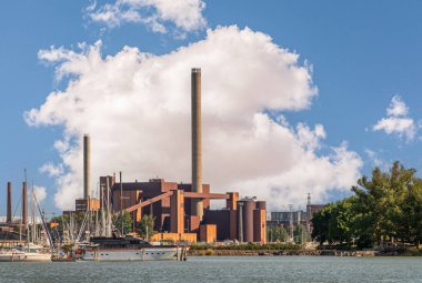 Helsinki, Finlandiya - 20 Temmuz 2022: Pohjoissatama Limanı. Kırmızı taş Hanasaari Enerji Santrali. Mavi bulutlara karşı uzun bacaları var. Liman suyu ve sone yeşili yapraklarda bir gemi.