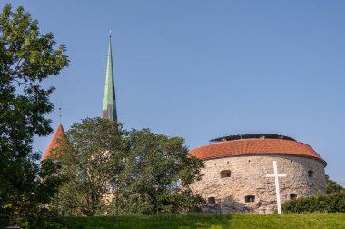 Estonya, Tallinn - 21 Temmuz 2022: Eski şehir, beyaz kule ve St. Olaf Kilisesi 'nin yeşil kulesi, bej rengi taştan yapılmış kuzey Suur Rannavarav kale kulesinin arkasında mavi gökyüzüne karşı. Biraz yeşil yaprak.