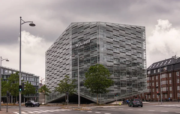 stock image Copenhagen, Denmark - July 23, 2022: Modern gray, glass and steel Nykredit bank building at corner of Puggaardsgade and Kalvebod Brygge under gray cloudscape. Cars, foliage, and buildings