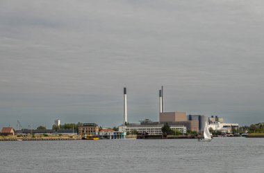 Kopenhag, Danimarka - 24 Temmuz 2022: Limanın doğu yakasında gri gökyüzü, bacalar ve binalar altında enerji üretim ve su arıtma tesisi, su üzerinde yelkenli yat