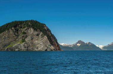 Diriliş Körfezi, Alaska, ABD - 22 Temmuz 2011: manzara. Yeşil ormanlık gri kaya, mavi gökyüzünün altında mavi okyanus suyunda bulunur. Ufukta kar parçaları olan dağ sırası