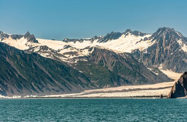 Diriltme Körfezi, Alaska, ABD - 22 Temmuz 2011: Buzulun mavi gökyüzünün altındaki koy suyuna ulaştığı körfez manzarasının batı tarafı. Koyu kayalık sıradağlarda bolca kar var.