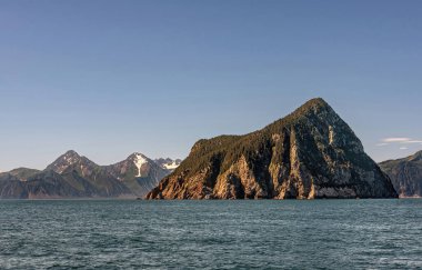 Diriliş Körfezi, Alaska, ABD - 22 Temmuz 2011: Açık mavi gökyüzünün altında mavi okyanus suyunda 1 izole edilmiş yeşil ormanımsı Hive Adası ile manzara. Ufukta karla kaplı dağlar...