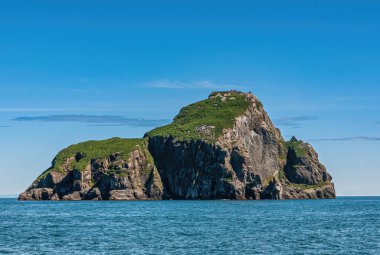 Resurrection Bay, Alaska, USA - July 22, 2011: Closeup of Hive Island, triangular rocky and partly covered by green vegetation in front of endless blue ocean under blue cloudscape clipart