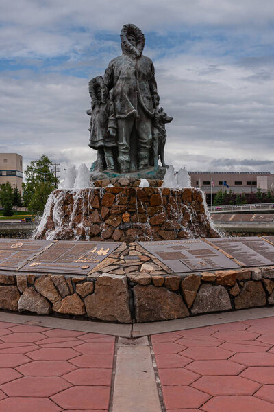 Фэрбанкс, Аляска, США - 27 июля 2011 года: Неизвестная статуя Первой Семьи и тупик. Крупный план отца с ребенком и собакой под голубым облаком. Таблички с текстом внизу. Флаги на расстоянии