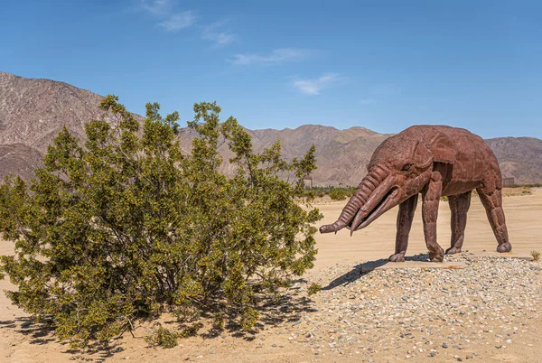 Borrego Springs, CA, ABD - 24 Nisan 2023: Kahverengi paslı metal, tarih öncesi dev karıncayiyen mamut heykeli kumlu çöl tabanına kurulmuş ve mavi gökyüzünün altında yeşil çalılar var. Arka taraftaki dağ sırası.