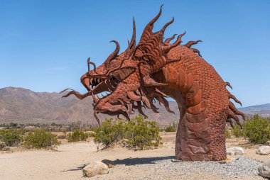 Borrego Springs, CA, ABD - 24 Nisan 2023: Kahverengi paslı metal, tarih öncesi dev yılan dinozor heykeli, yandan kumlu çölde mavi gökyüzü altında, yeşil çalılıklar, dağ sıraları