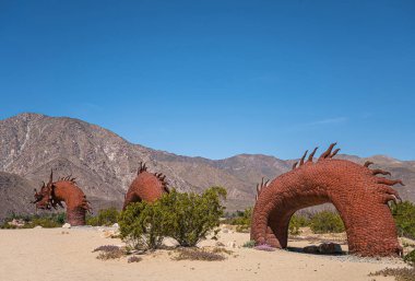 Borrego Springs, CA, ABD - 24 Nisan 2023: Kahverengi paslı metal, tarih öncesi dev yılan dinozor heykeli, 3 ön parça, kumlu çölde mavi gökyüzü altında, yeşil çalılıklar, dağ sıraları