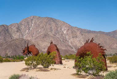 Borrego Springs, CA, ABD - 24 Nisan 2023: Kahverengi paslı metal, tarih öncesi dev yılan dinozor heykeli, ön kısımları, kumlu çölde mavi gökyüzü altında, yeşil çalılıklar, dağ sıraları