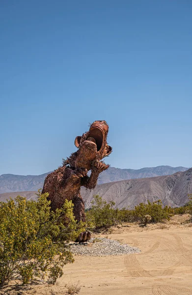 Borrego Springs, CA, ABD - 24 Nisan 2023: Mavi gökyüzü, yeşil çalılıklar, dağ sıraları altında kumlu çöl üzerinde kahverengi paslanmış tarih öncesi dev uluyan dinozor heykeli ön görünümü