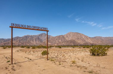 Borrego Springs, CA, ABD - 24 Nisan 2023: Un diyalogu, dos sohbet kemeri mavi gökyüzünün altında ve ufukta kahverengi taş sıradağları var. Bazı yeşil çalılar