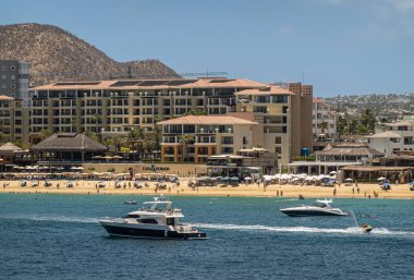 Meksika, Cabo San Lucas - 16 Temmuz 2023 Casa Dorado, Blues gökyüzünün altındaki mavi su koyunda sarı plaj cephesi. Kum üzerindeki şemsiye çizgileri ve eğlence tekneleri ve suda scooterlar.