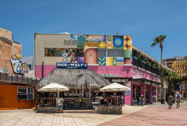 Mexico, Cabo San Lucas - July 16, 2023: Colorfull architecture and signs of Mayan Monkey hostel, a pharmacy and bars in Marina Mercado under blue sky. Shoppers in alley clipart