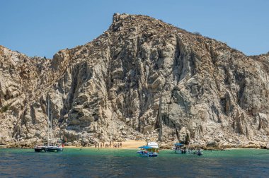 Meksika, Cabo San Lucas - 16 Temmuz 2023: küçük plaj, bol banyo, arazi yakınlarındaki yüksek gri kayalıkların altında yeşil körfez suyunda birkaç küçük tekne. Açık mavi gökyüzü