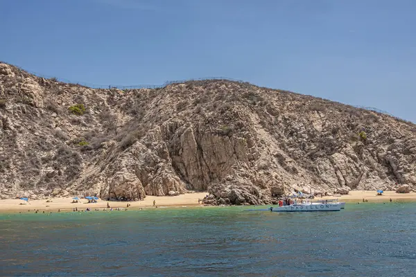 Meksika, Cabo San Lucas - 16 Temmuz 2023: kıyı şeridindeki bej-kahverengi kayalık tepeler. Papillon katamaranı körfez suyundaki turist gemisi olarak. Kumsaldaki ve sörfteki insanlar