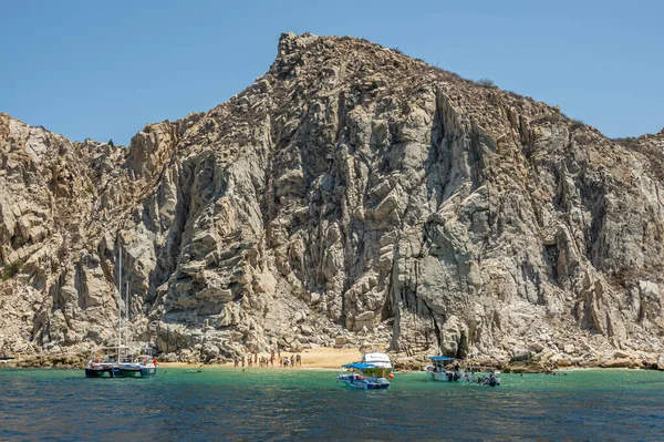 Meksika, Cabo San Lucas - 16 Temmuz 2023: küçük plaj, bol banyo, arazi yakınlarındaki yüksek gri kayalıkların altında yeşil körfez suyunda birkaç küçük tekne. Açık mavi gökyüzü